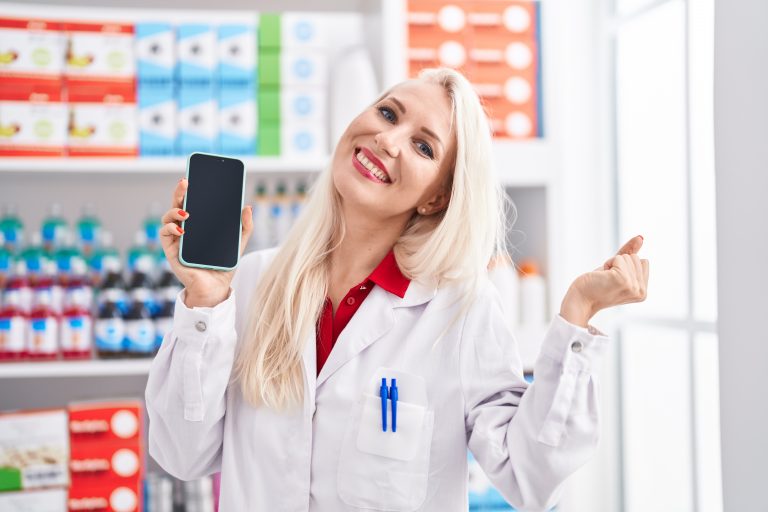 Caucasian woman working at pharmacy drugstore showing smartphone screen screaming proud, celebrating victory and success very excited with raised arms