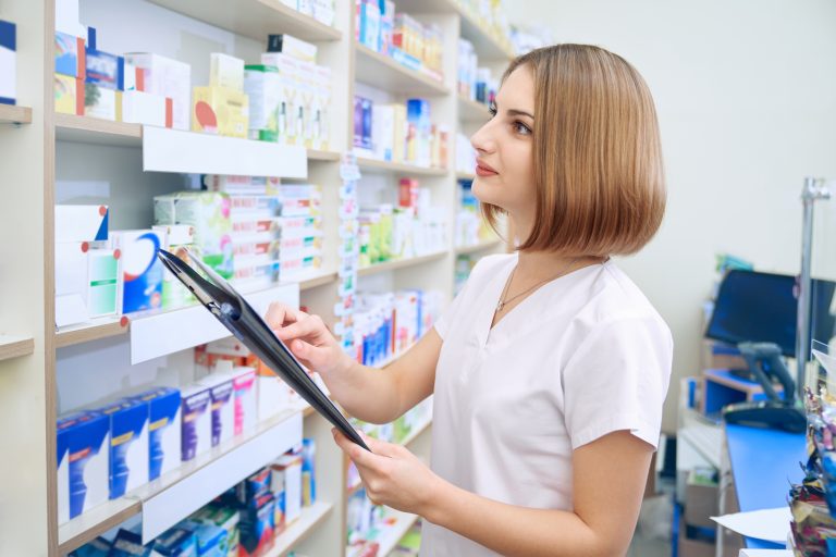 Female pharmacist standing near shelves with medicaments in drugstore. Side view of woman in white lab coat holding folder in hands. Specialist working in pharmaceutical store. Medicine concept.